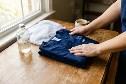 Hands folding custom scrub top on wooden table with detergent and mesh laundry bag